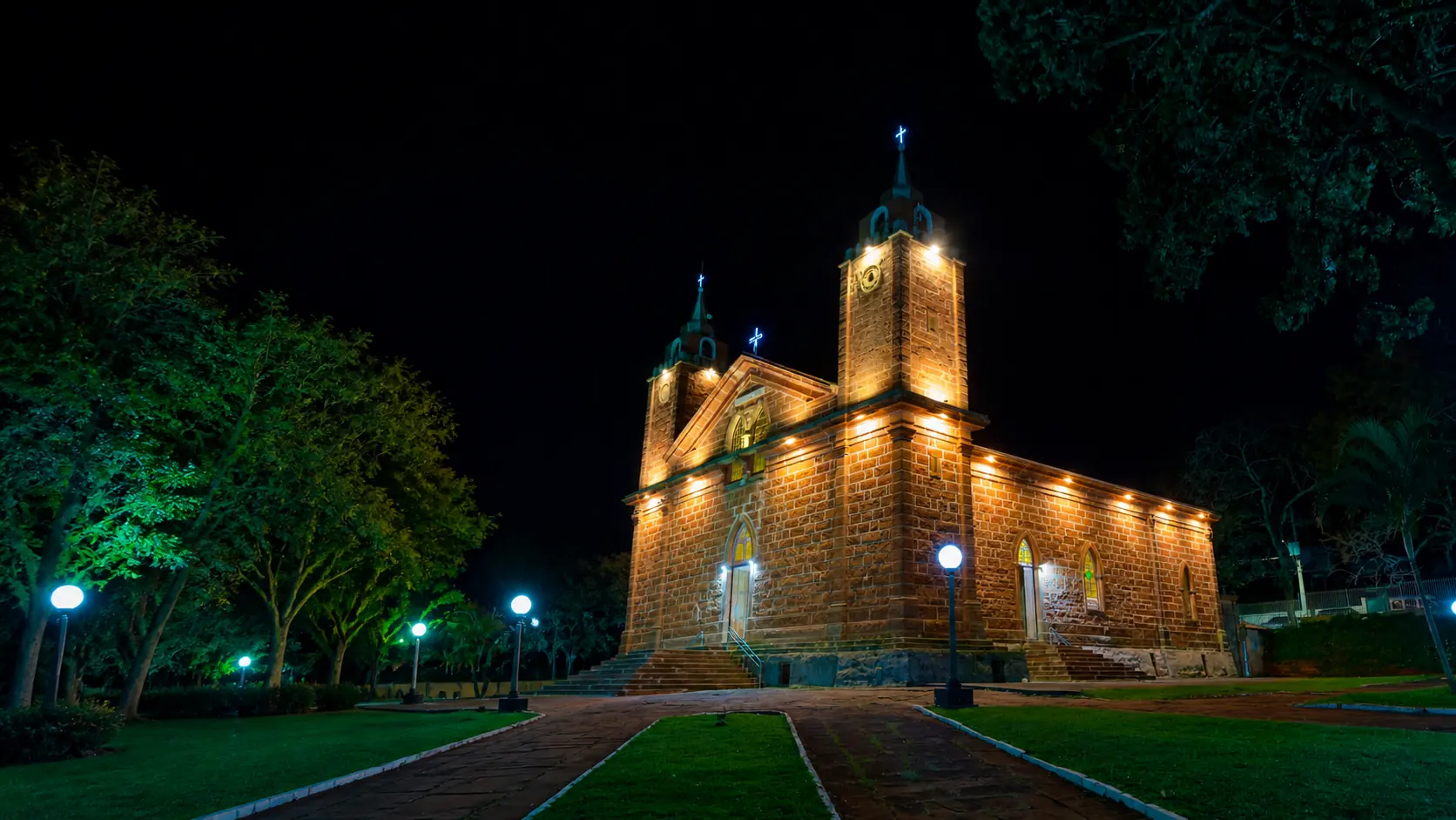 Igreja histórica iluminada à noite por refletores, com a praça e o caminho iluminados por postes de LED